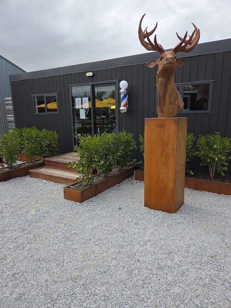 The Highway Barbers building on Main Road, Luggate: black vertical cladding, a gravel forecourt, a barber pole beside the door and a rusted steel stag on a plinth.