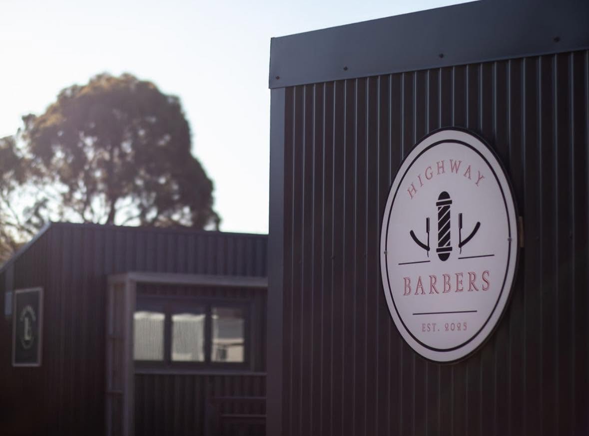 The Highway Barbers roundel sign on the shop's black corrugated cladding at Luggate, gum tree behind it in evening light.